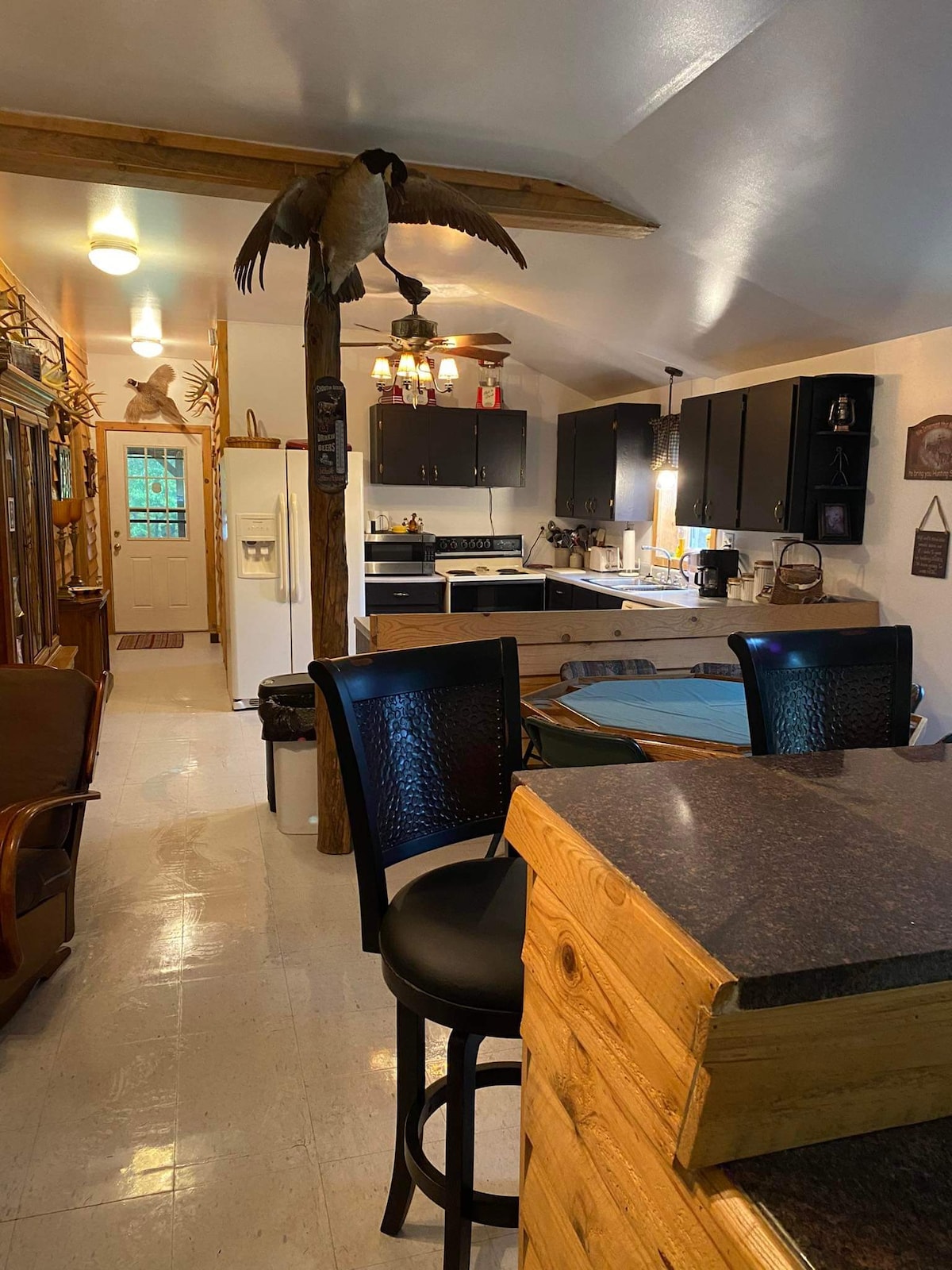A well-appointed kitchen features dark cabinetry and modern appliances, with a dining area visible in the foreground. A rustic island with bar seating offers additional space. Natural light enters through a nearby window, enhancing the open layout and warm ambiance of the living space.