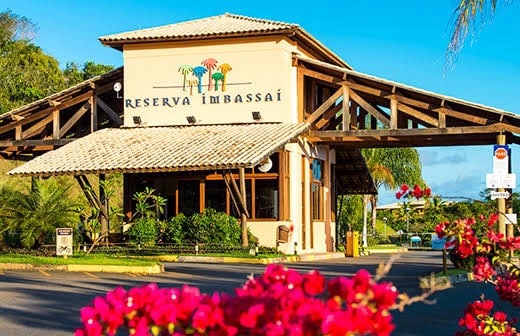 The entrance to Reserva Imbassai is framed by vibrant flowers and lush greenery. The wooden structure showcases a welcoming design, with a sloped roof and large overhangs. A sign prominently displays the name, inviting guests to explore the amenities of this upscale condominium.