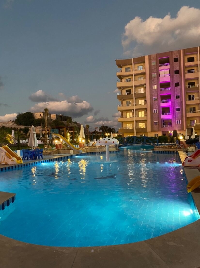 A large outdoor pool is illuminated with soft blue lights, reflecting the evening sky. Surrounding the pool are bright umbrellas and seating areas. The building facade in the background features colorful lighting, enhancing the tranquil ambiance of the resort.