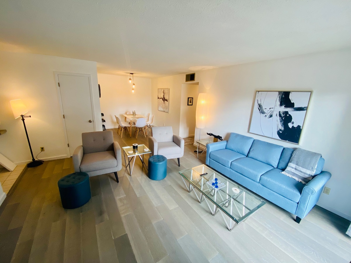A living area is presented with a blue sofa and two armchairs positioned around a glass coffee table. Soft lighting from a floor lamp enhances the room's illumination. The dining area is visible in the background, featuring a simple table and chairs.