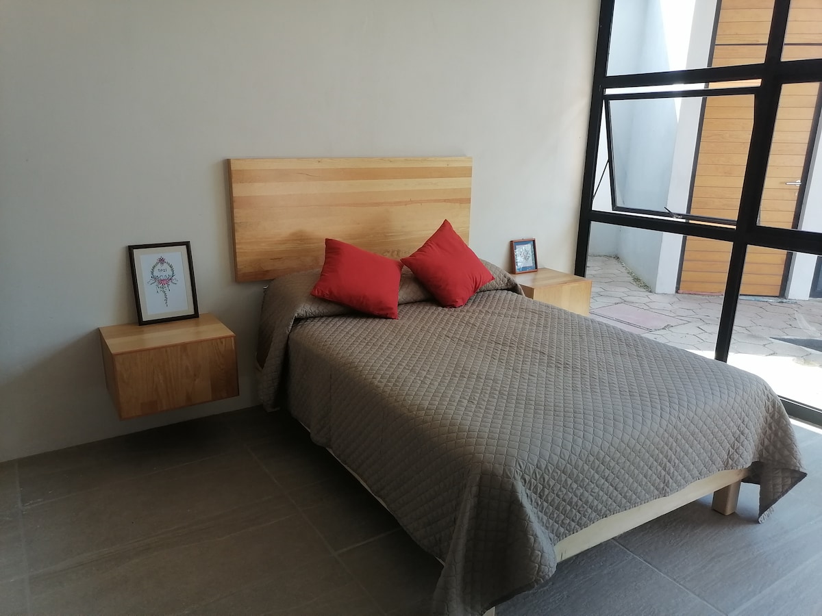 A spacious bed with a neutral quilted cover is positioned against a simple wooden headboard. Two red accent pillows add a pop of color. Nightstands with framed artwork sit on either side, while large glass windows flood the room with natural light.