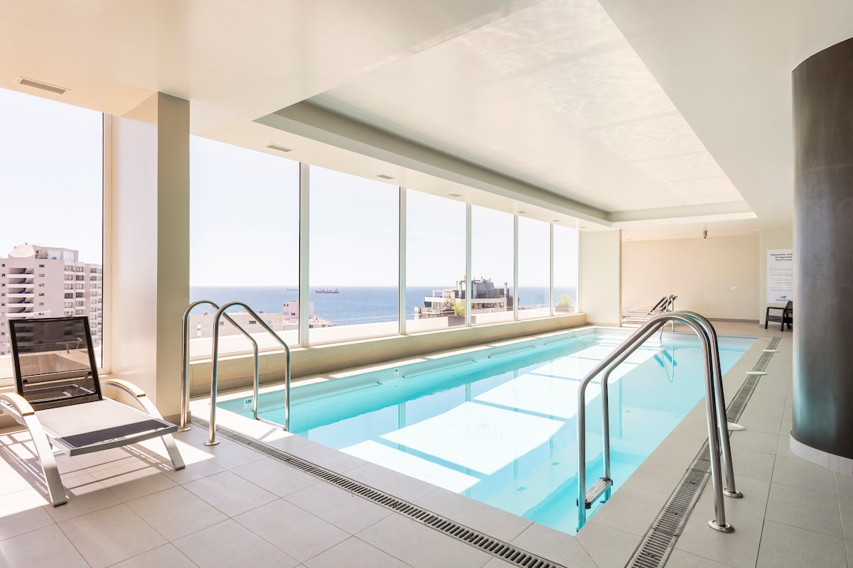 An inviting indoor pool is framed by large windows that offer views of the ocean and cityscape. The area is designed with minimalist decor, featuring a sleek lounge chair positioned beside the water, and natural light floods the space, enhancing its open feel.