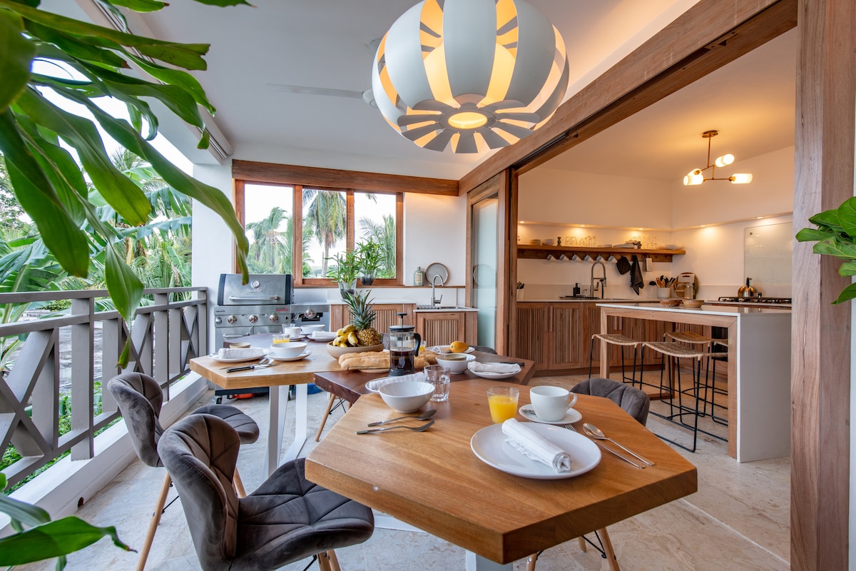 An integrated dining area features a wooden table set for breakfast, complemented by stylish chairs. Large windows allow natural light to illuminate the space, with a modern light fixture overhead. A glimpse of the outdoor kitchen area with a gas grill is visible in the background.