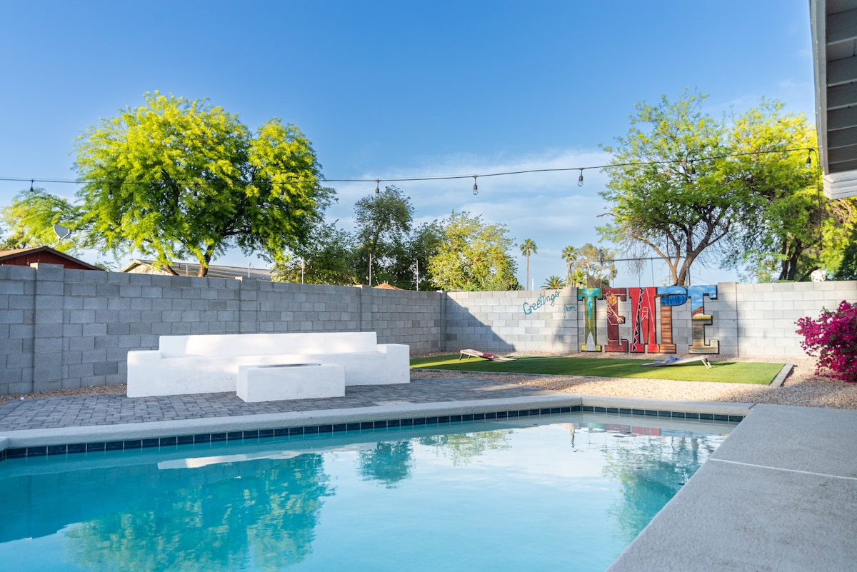 A spacious backyard features a heated pool with clear water, surrounded by a concrete patio. Colorful letters spelling 'TEMPE' adorn the wall, accompanied by vibrant greenery and landscaping. Soft seating is arranged near the pool, offering a relaxed outdoor space.