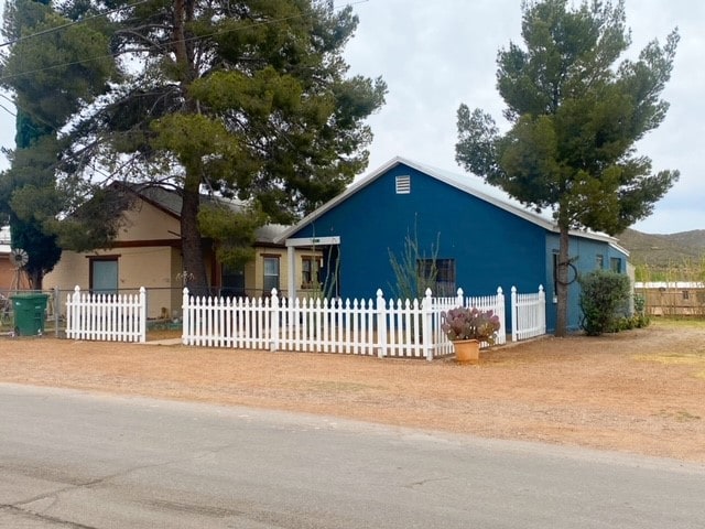 A two-bedroom home is surrounded by a white picket fence, featuring a blue exterior and a sloped roof. A gravel pathway leads to the entrance, which is flanked by tall pine trees. The yard includes a potted plant visible near the entrance.