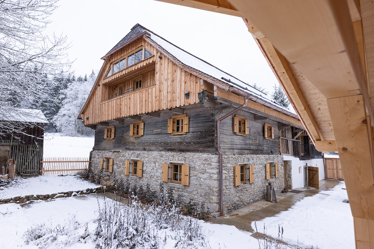 The historical timber-framed house features a combination of stone and wooden elements, with several large windows adorned with wooden shutters. Snow covers the surrounding landscape, enhancing the house's rustic charm. A gravel path leads to the entrance, surrounded by a serene winter scene.