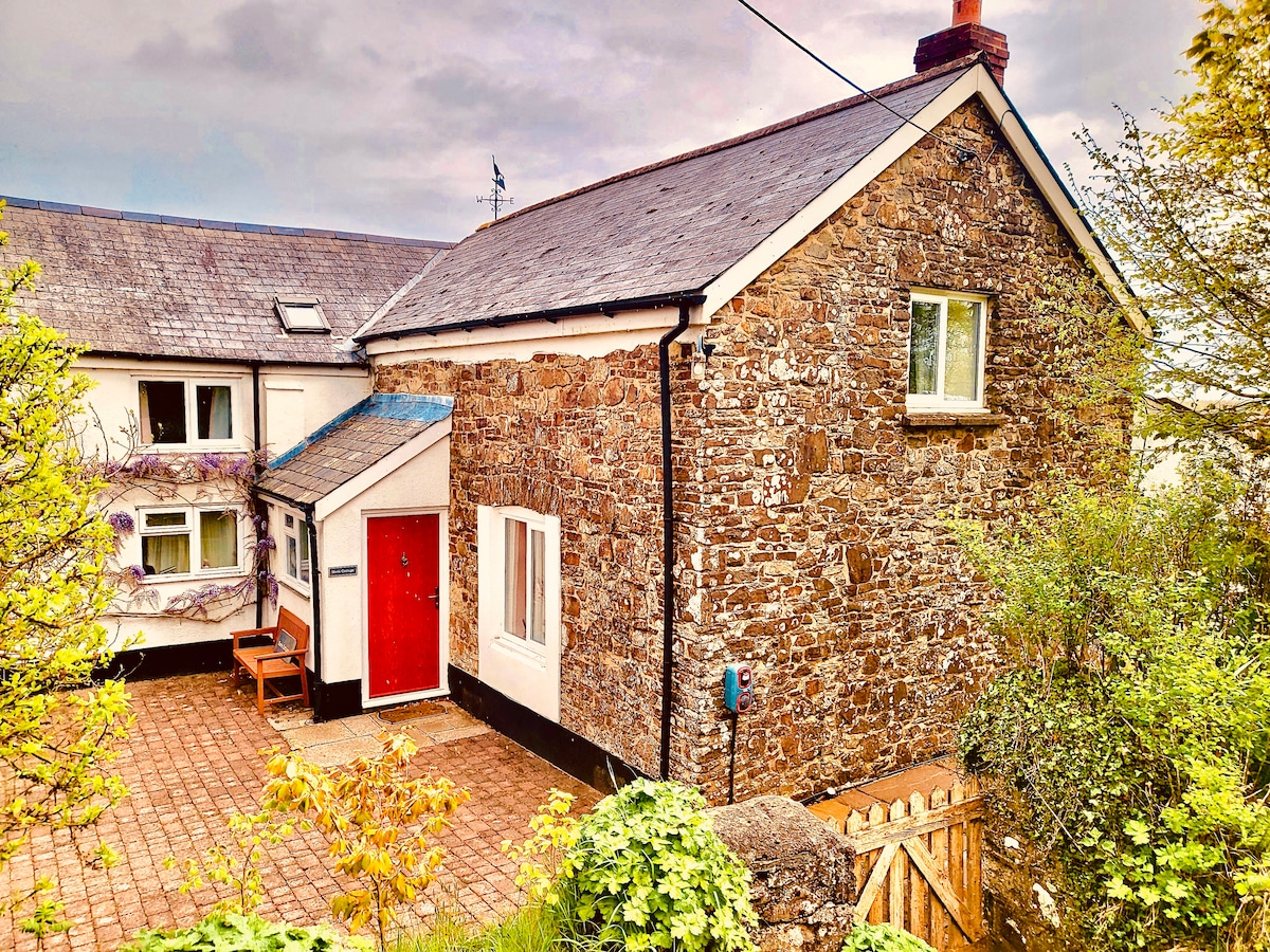 The exterior of the stone cottage features a welcoming red door and adjacent seating area. Lush greenery surrounds the property, complemented by a paved pathway leading to the entrance. The structure showcases a blend of rustic charm and modern living.