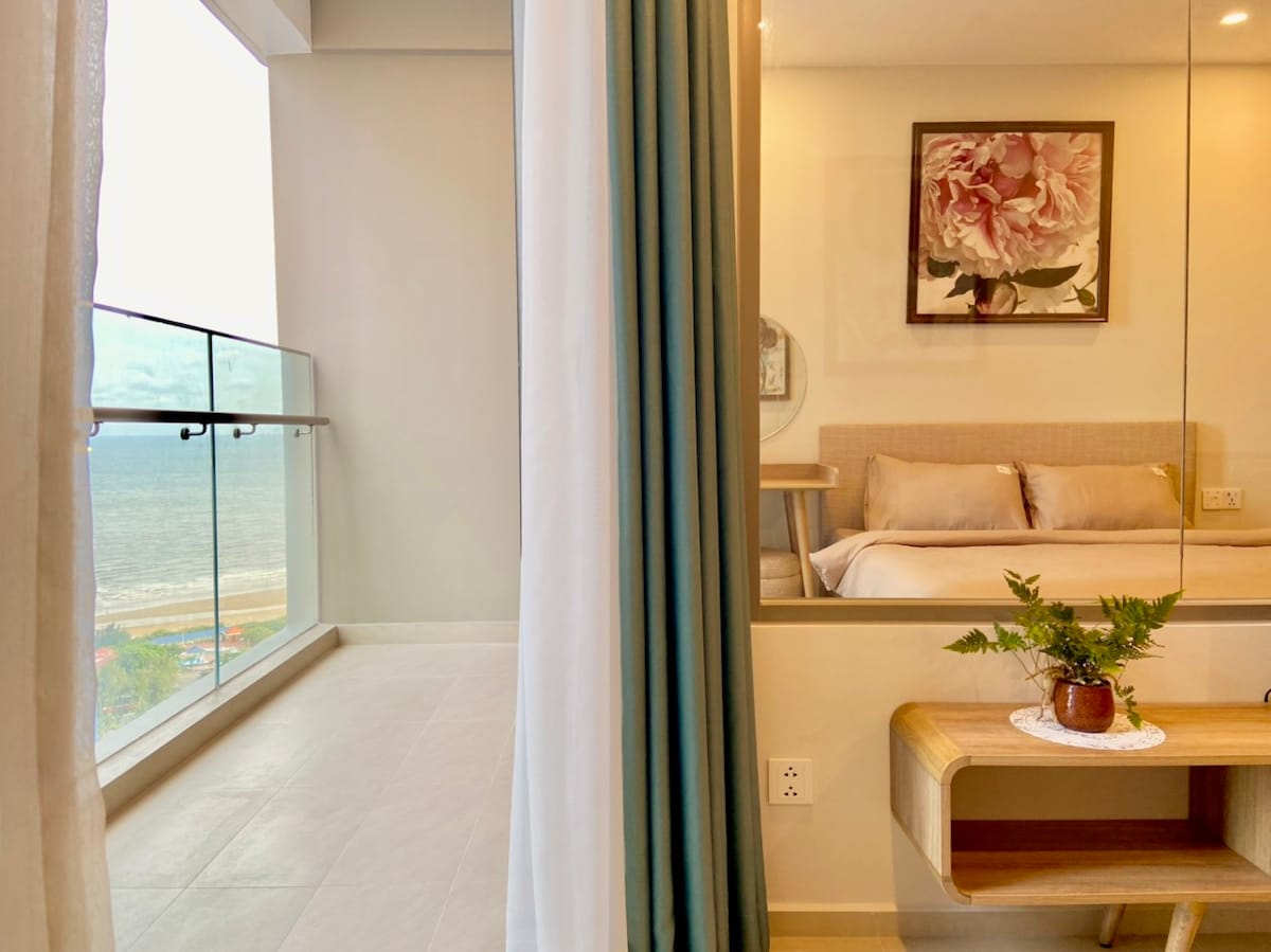 A spacious interior features a comfortable bed with neutral-colored bedding, complemented by a decorative wall art piece. A large window allows natural light to illuminate the room, while a small table with a plant adds a touch of greenery. A balcony with a sea view is visible, enhancing the sense of openness.
