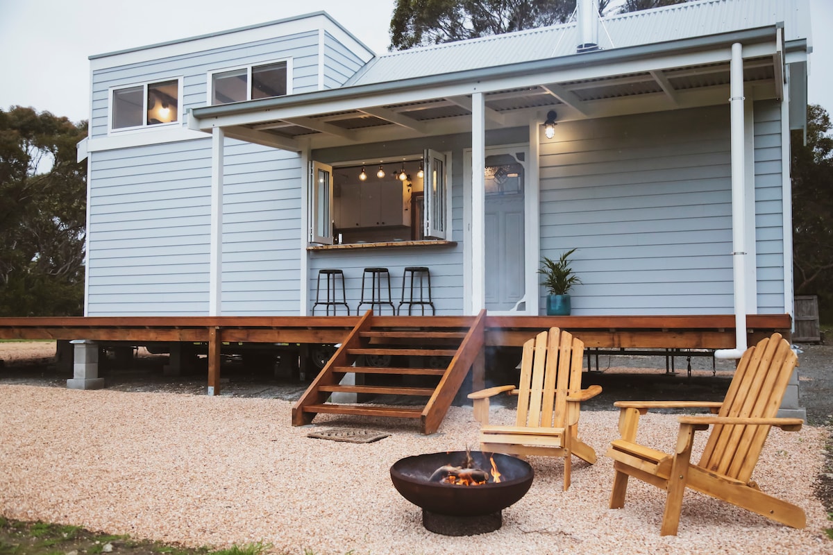 A charming tiny home is nestled among the gum trees, featuring a spacious wooden deck with two adirondack chairs positioned near a fire pit. Large windows invite natural light, while a well-equipped indoor kitchen is visible through the open door.