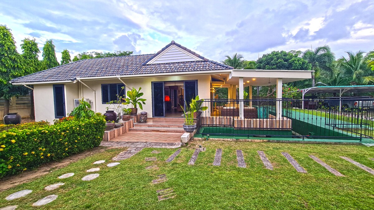 A charming villa is depicted with a sloped roof and light-colored exterior. Lush greenery surrounds the property, while a wooden deck leads to an outdoor area. Potted plants and decorative elements enhance the entryway, with a glimpse of a secure terrace in the background.