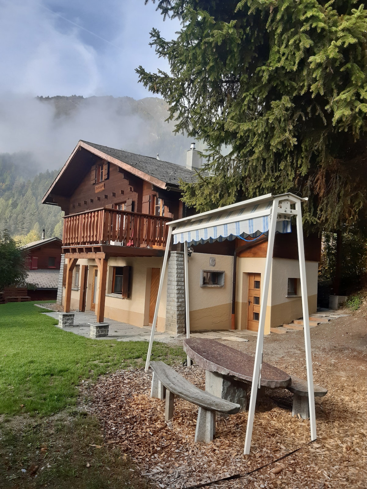 A charming chalet exterior is framed by trees and mountains, highlighting a grassy area. A wooden swing sits near a stone table surrounded by benches. The house features a large balcony and a welcoming entrance, complemented by a serene atmosphere.