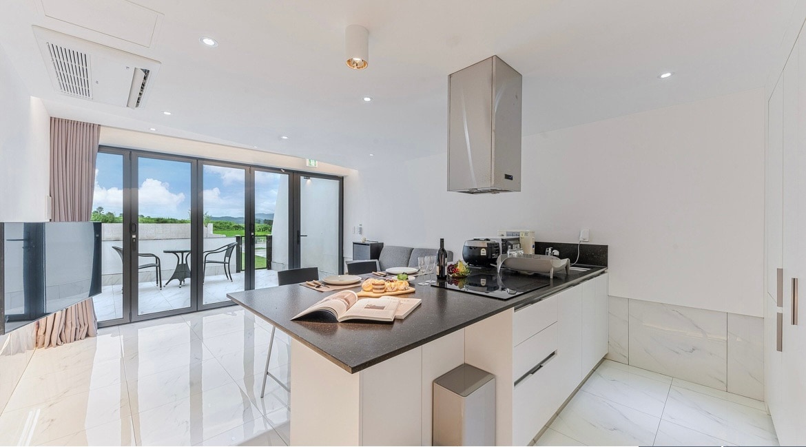 A modern kitchen with sleek cabinetry and a central island is featured. Large glass doors open to a patio, inviting natural light. A cooking area with stainless steel appliances and a countertop is visible, along with a neatly arranged cookbook and pastries on display.