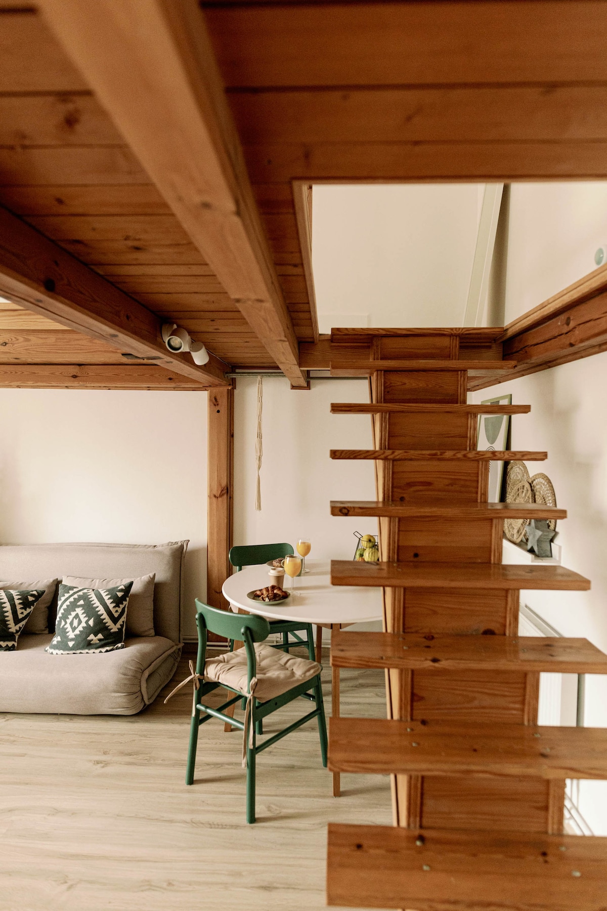 A cozy living area features a gray sofa adorned with patterned cushions. A wooden staircase leads to a lofted space. A simple dining table with green chairs is set nearby, creating a warm and welcoming environment.