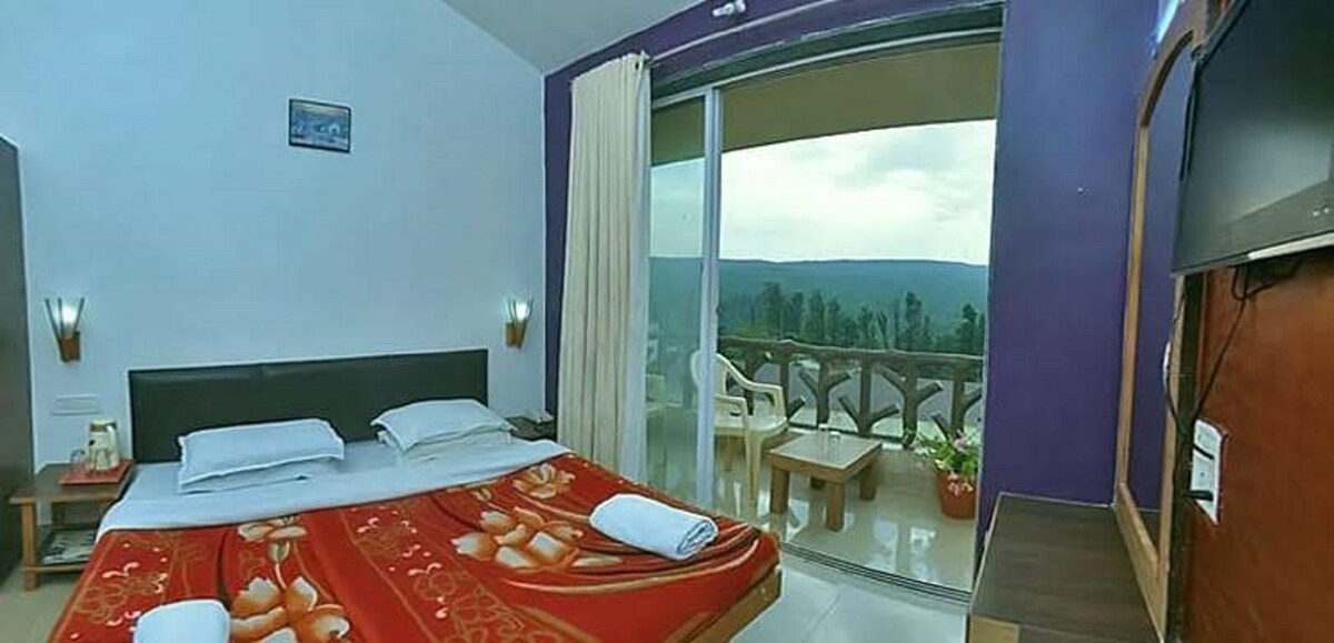 A guest room features a comfortable bed adorned with a red blanket. Large glass doors open to a balcony with mountain views. Natural light fills the space, complemented by a wall-mounted television and a small seating area with white chairs. Soft curtains frame the window.