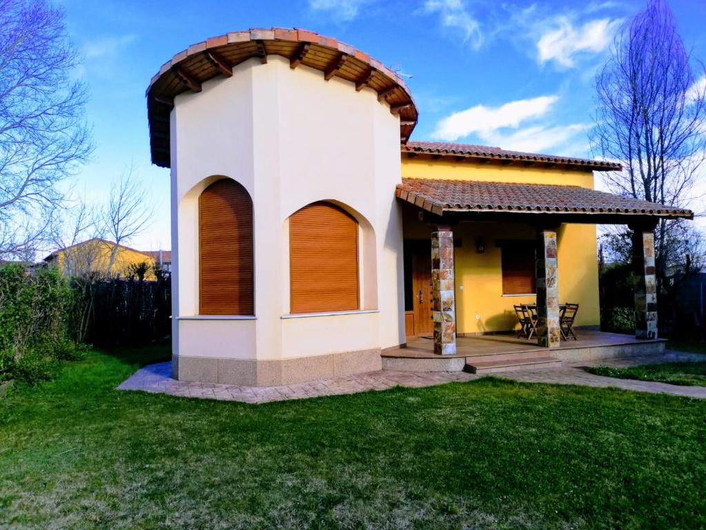 A charming single-family home is depicted, featuring a circular design with a tiled roof. Large arched windows are adorned with wooden shutters. A covered porch offers a small outdoor seating area surrounded by a well-maintained lawn and a backdrop of trees.
