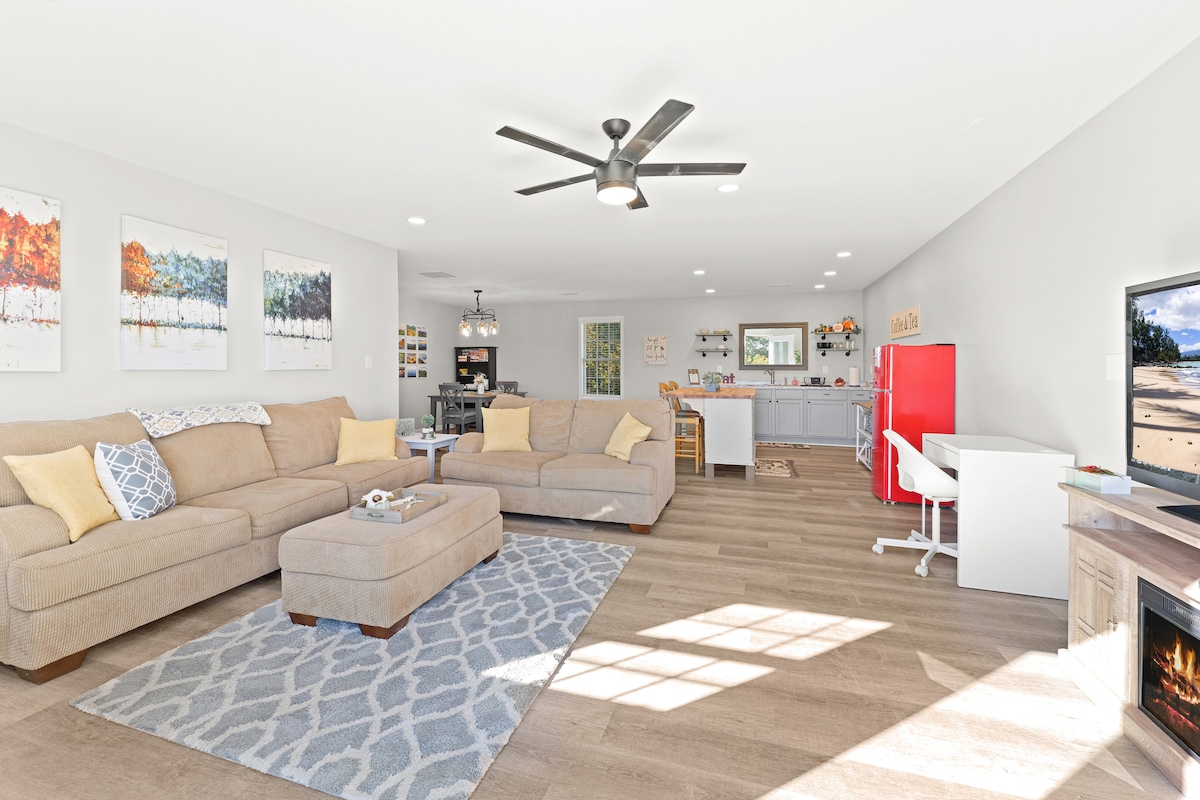 The spacious living area features a light-colored sectional sofa arranged around a central ottoman. A large area rug adds warmth to the space, complemented by a modern ceiling fan. A cozy entertainment unit with a television is visible, and a kitchenette can be seen in the background.
