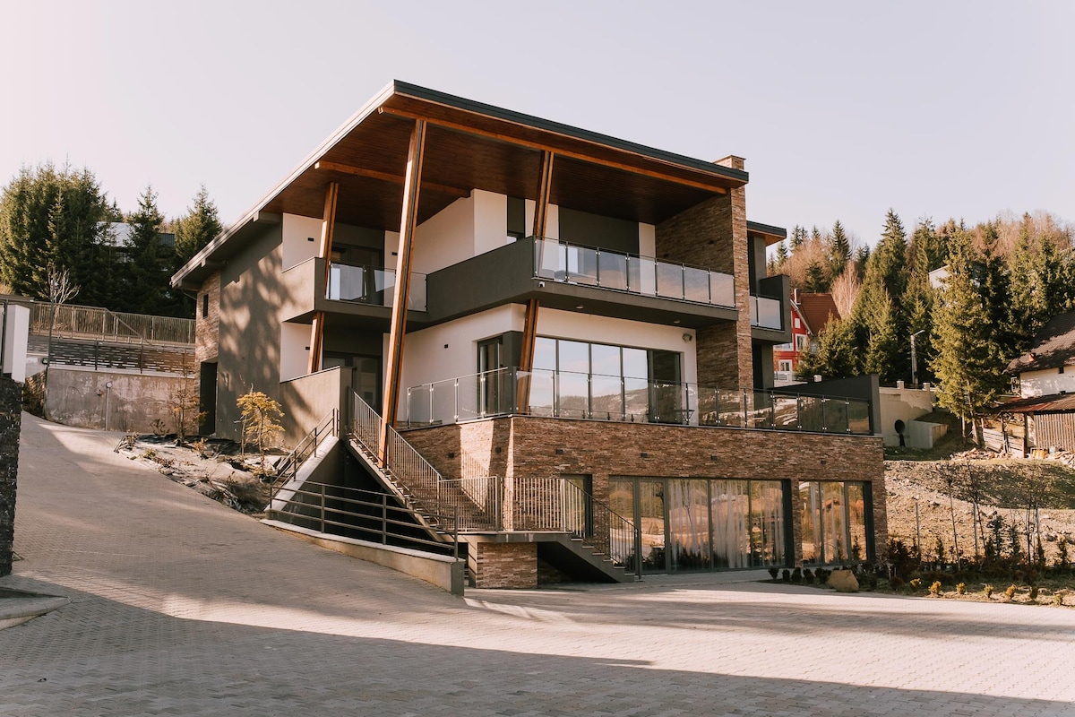 A modern building with a two-story design features large glass windows and a spacious balcony. The stone and wood exterior blends with the surrounding landscape. A landscaped area is visible in the foreground, serving as a welcoming entrance to the property.