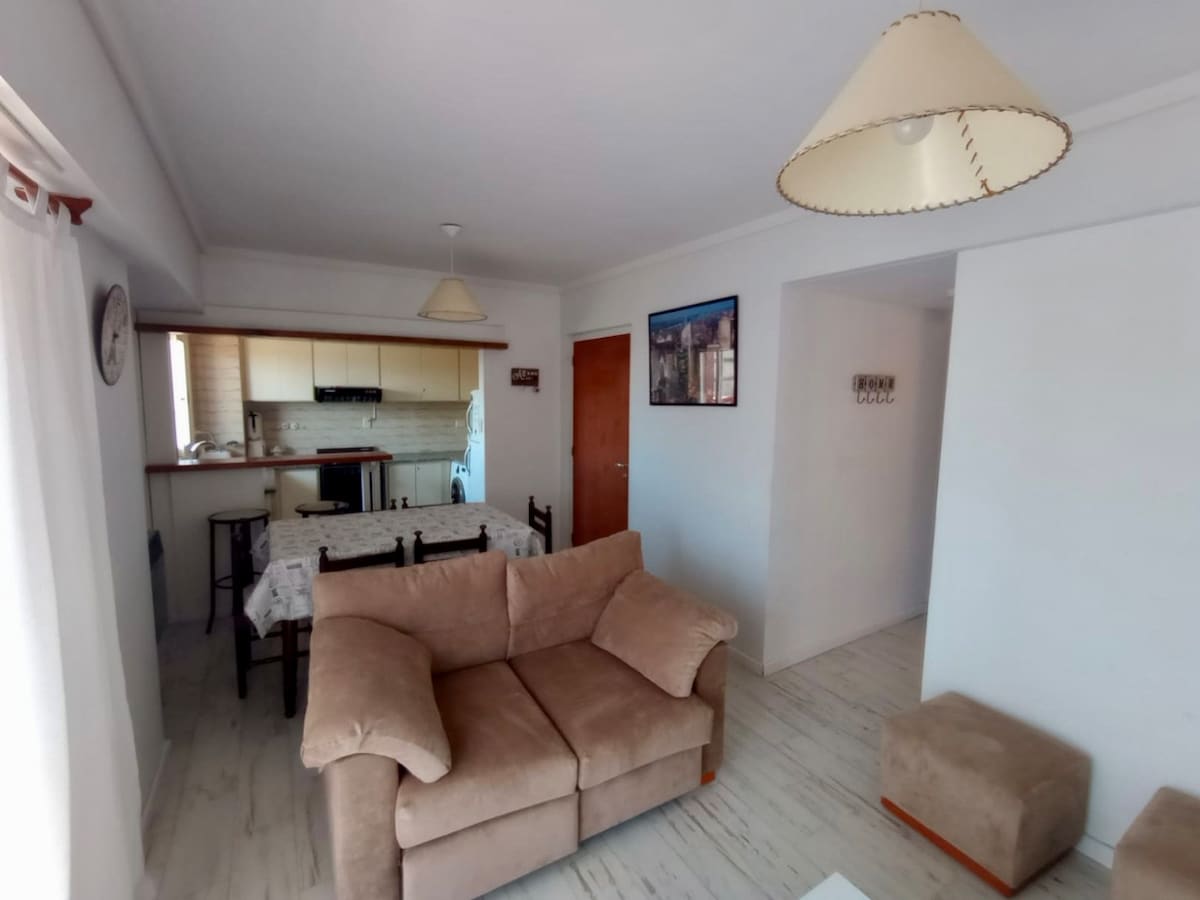 A living space features a comfortable beige sofa and a small ottoman, arranged around a light-colored coffee table. A dining area is adjacent, showcasing a table set for four. The room is illuminated by two pendant lamps and includes an open view towards the kitchen.