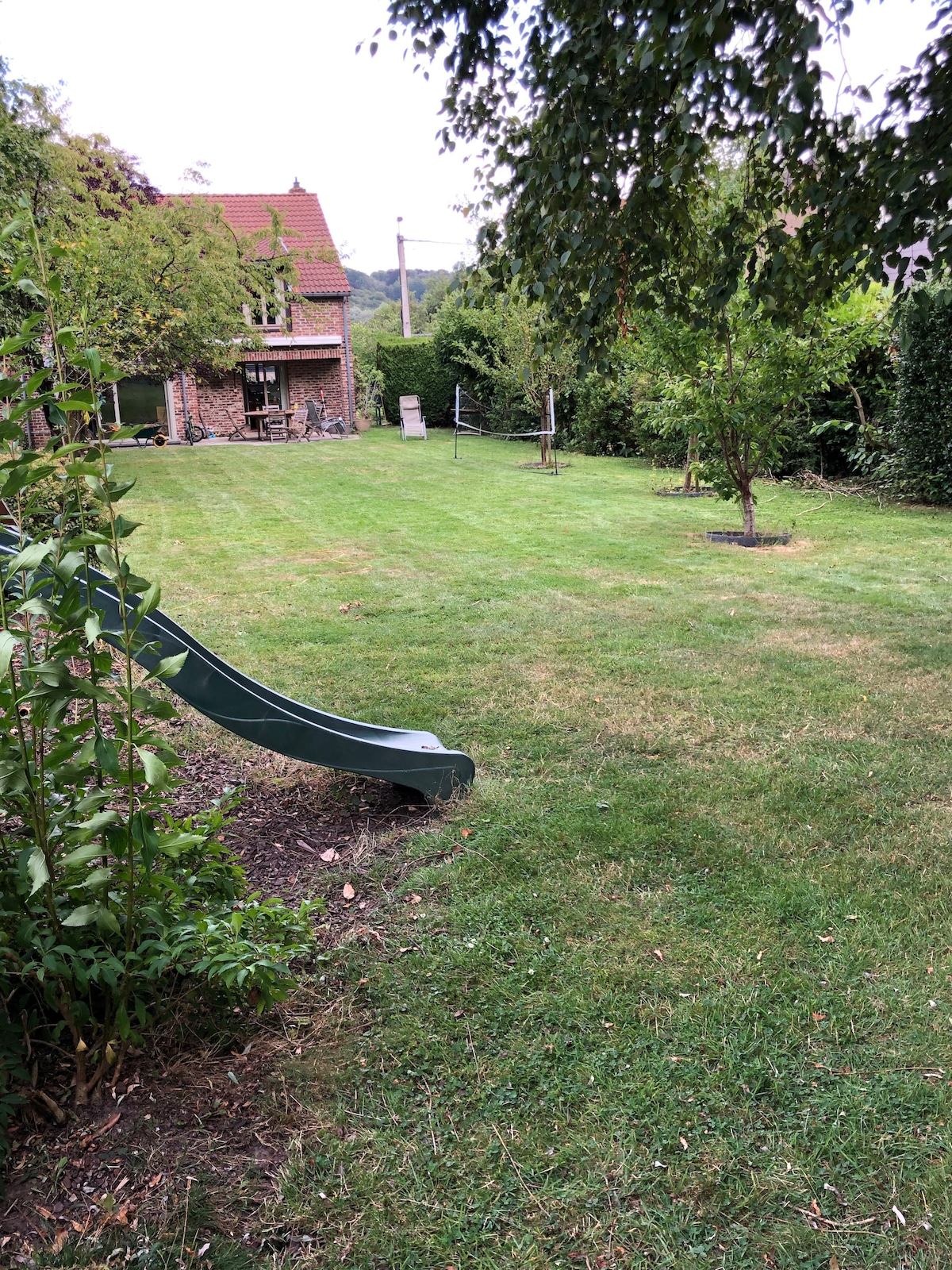 A spacious garden is shown with a grassy lawn, featuring a green slide on the left side. Small trees and shrubs border the area, while a home is visible in the background. The setting is calm and well-maintained, ideal for outdoor activities.