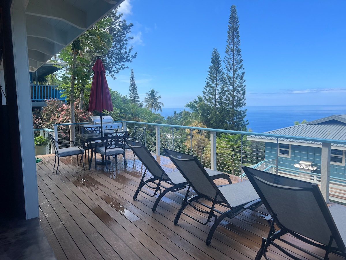 A private deck features a dining table with six chairs and four lounge chairs, inviting relaxation. A red umbrella provides shade, while the expansive ocean view showcases clear blue skies and lush greenery. The wooden flooring maintains a natural aesthetic.