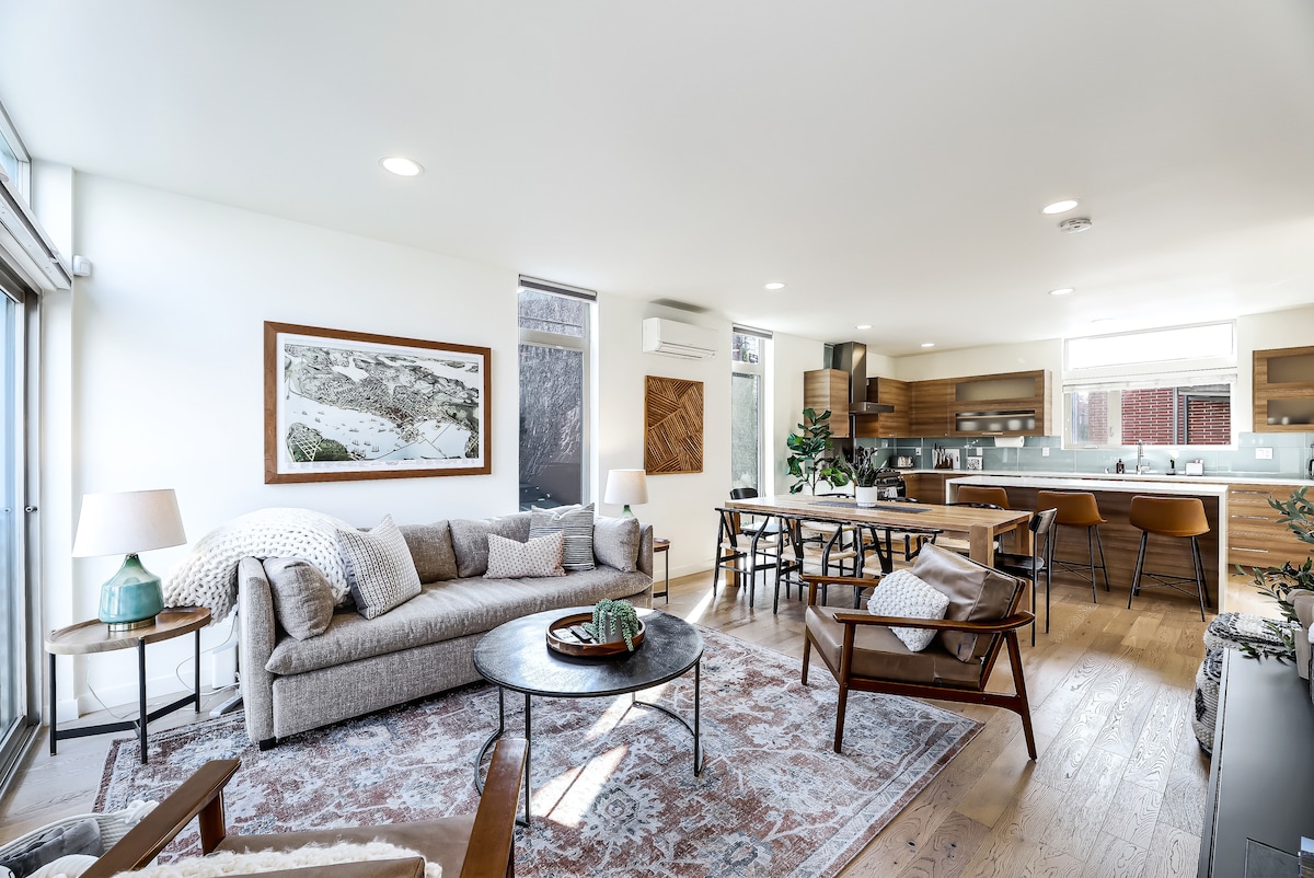 A modern living area is depicted with a large, comfortable sofa and two armchairs arranged around a circular table. Natural light is provided through expansive windows. The open kitchen features wood cabinetry and a breakfast bar, complementing the hardwood floors throughout the space.