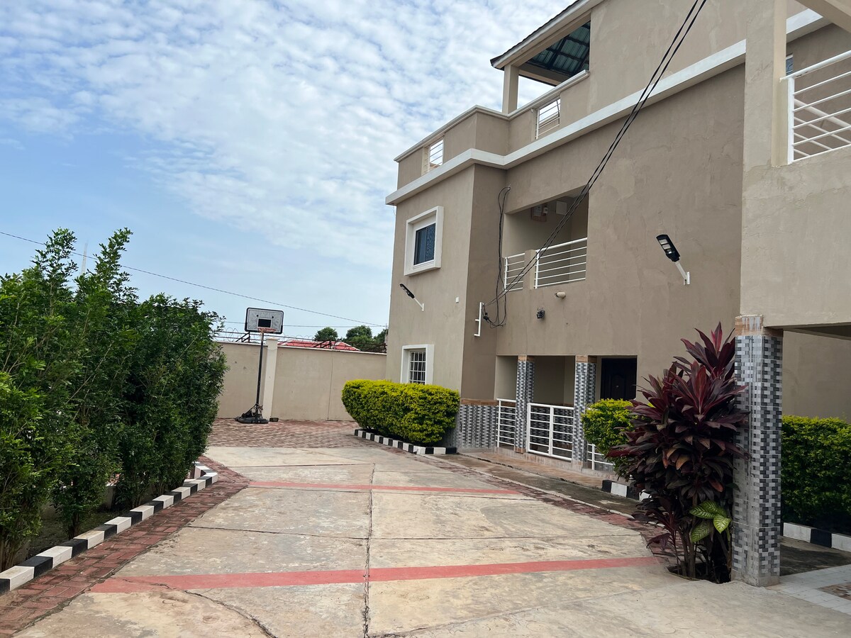 A spacious driveway leads up to a modern two-story building. Green shrubbery and decorative plants line the walkway. A basketball hoop is positioned near the side, contributing to the outdoor recreational space.