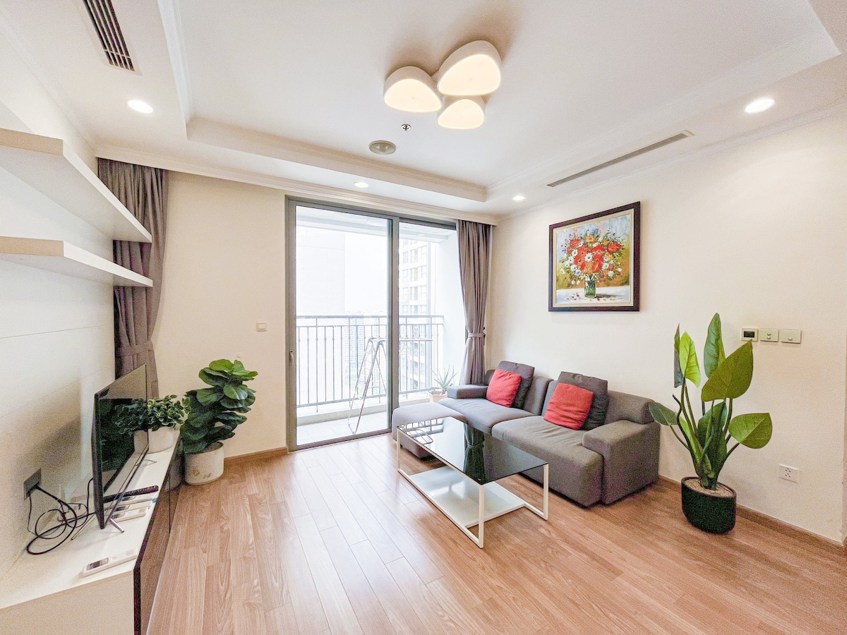 A modern living room is showcased with a comfortable gray sofa adorned with red cushions. A light wooden floor enhances the space, where a flat-screen TV is mounted on a sleek white shelf. A large window allows natural light to brighten the area, complemented by indoor plants.