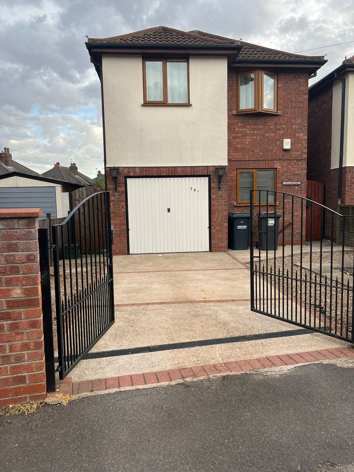 The home's exterior presents a two-story brick structure with a white garage door. A gated entrance leads to a spacious driveway. Surrounding elements include a neat lawn area and neighboring properties, framed against a cloudy sky.