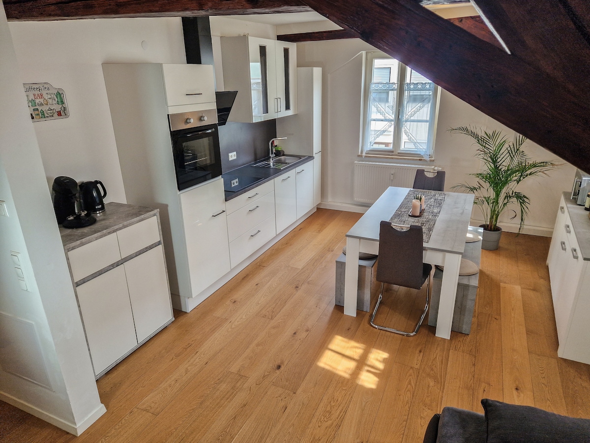 A modern kitchen area is showcased, featuring sleek white cabinets and a dark countertop. A dining table with gray chairs is positioned near a window, allowing natural light to illuminate the space. A potted plant adds a touch of greenery to the inviting environment.