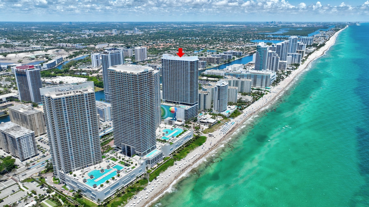 An aerial view captures the beachfront resort hotel surrounded by mid-rise and high-rise buildings along the coastline. Vibrant blue waters meet sandy shores, while the expansive skyline displays various architectural styles. The hotel is marked for easy identification.