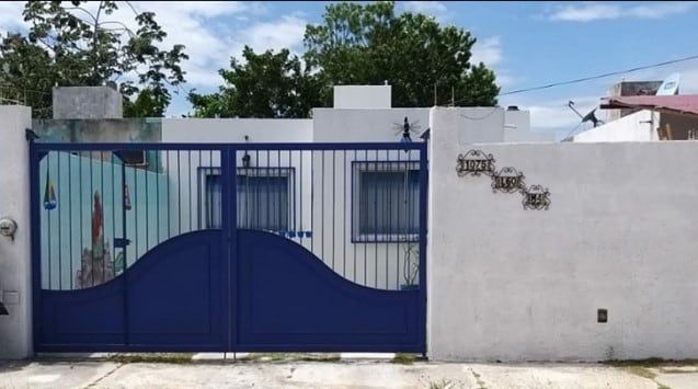 A blue metal gate is prominently displayed, providing security and privacy. The surrounding white walls contrast with the vibrant gate, while trees and foliage are visible in the background, creating a calm outdoor environment.