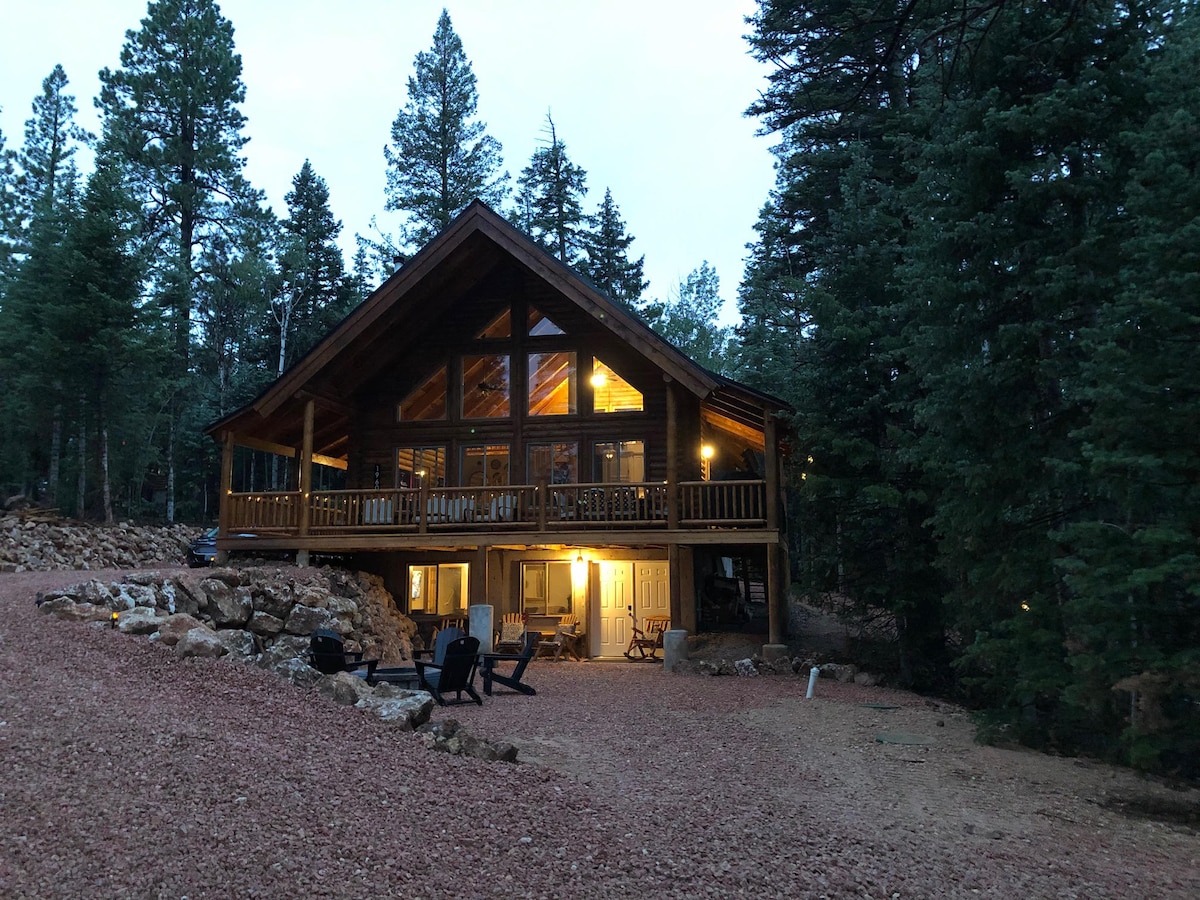 A modern rustic cabin is set among tall pine trees, featuring a large deck with multiple chairs. The structure showcases expansive windows, allowing natural light to fill the interior. A gravel pathway leads up to the front door, emphasizing the natural surroundings.