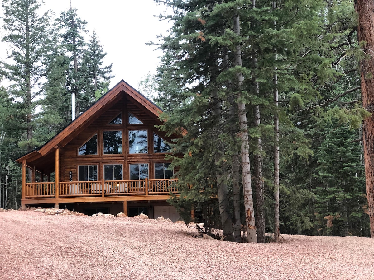 A modern rustic cabin is nestled among tall trees, featuring large glass windows that provide a view of the surrounding forest. A spacious deck is visible at the front, with wooden railings. Pebbled ground leads up to the cabin, blending into the natural landscape.