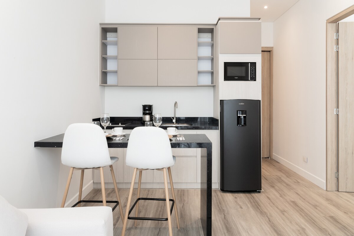 A modern kitchen area features a sleek black countertop with minimalist cabinetry above and below it. Two white chairs are positioned at a small dining table. A black refrigerator and microwave are visible, along with essential kitchen items arranged neatly.