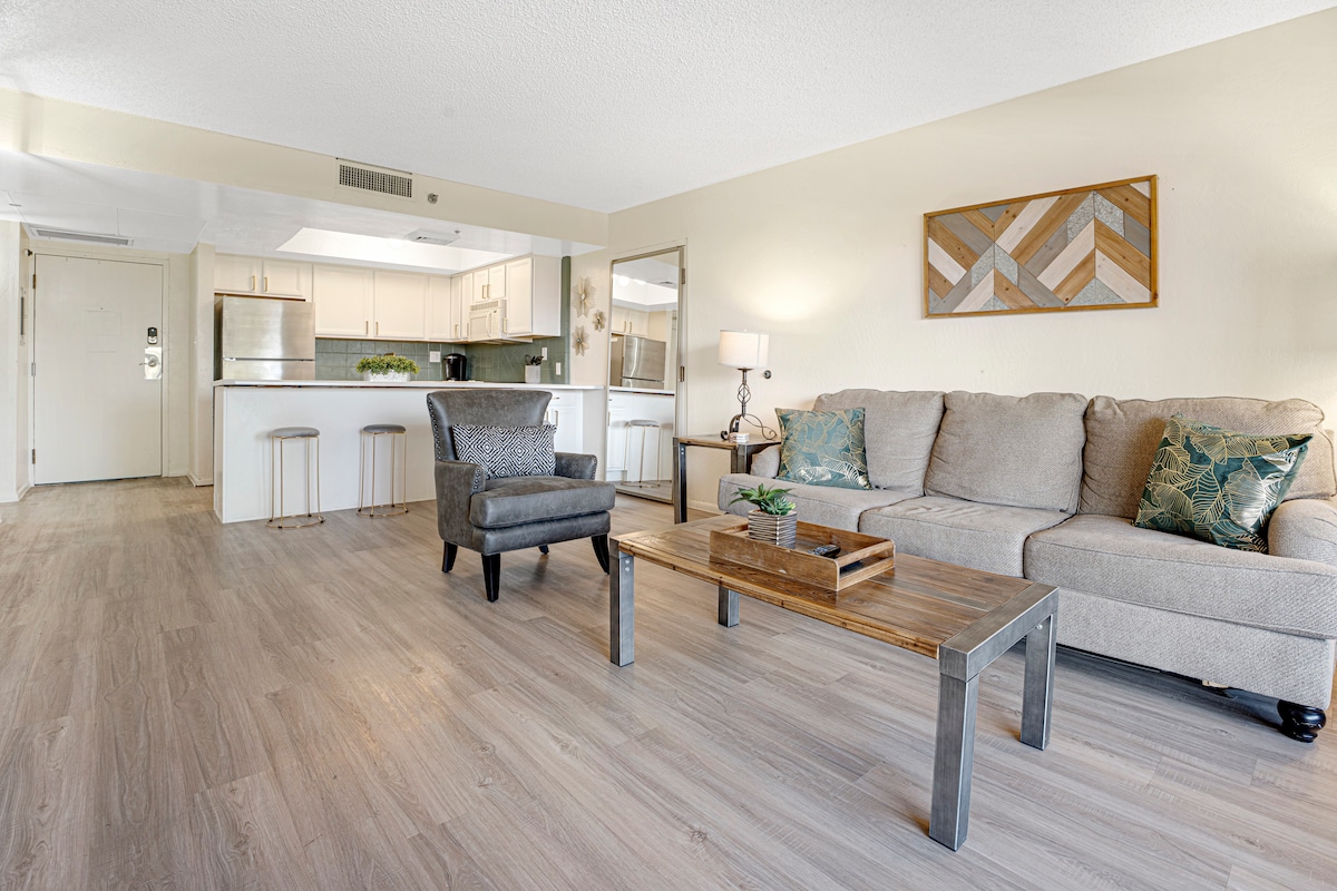 The living area features a cozy sofa with decorative cushions, a wooden coffee table with a small plant, and two bar stools positioned by the kitchen peninsula. Light-colored walls and a piece of wall art contribute to the bright, open atmosphere.