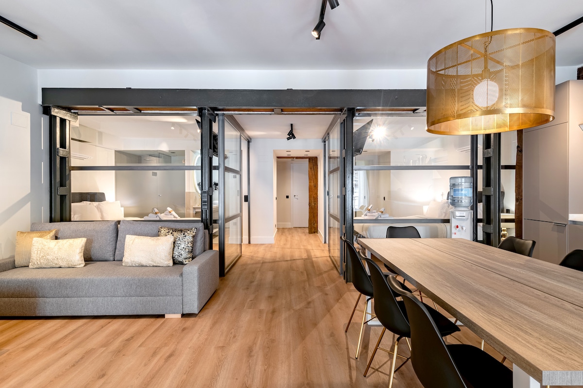 An open-concept living space features a modern gray sofa adorned with decorative cushions, paired with a spacious dining table. The room is illuminated by a stylish pendant light, with sliding glass doors leading to a well-equipped kitchen area, enhancing the overall lightness of the environment.