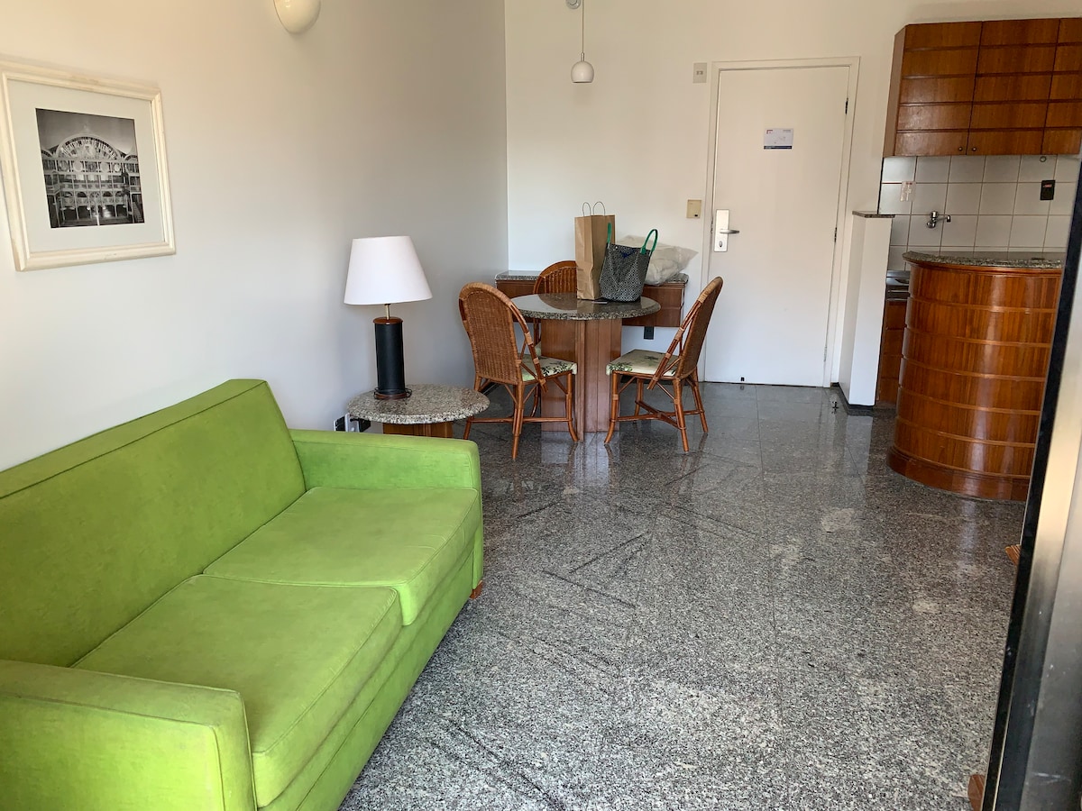 A green sofa is placed in the living area, accompanied by a round dining table with four wicker chairs. A modern lamp stands next to the sofa, and a small round table with a marble top is positioned nearby. The kitchen area features wood cabinetry and a tiled backsplash.