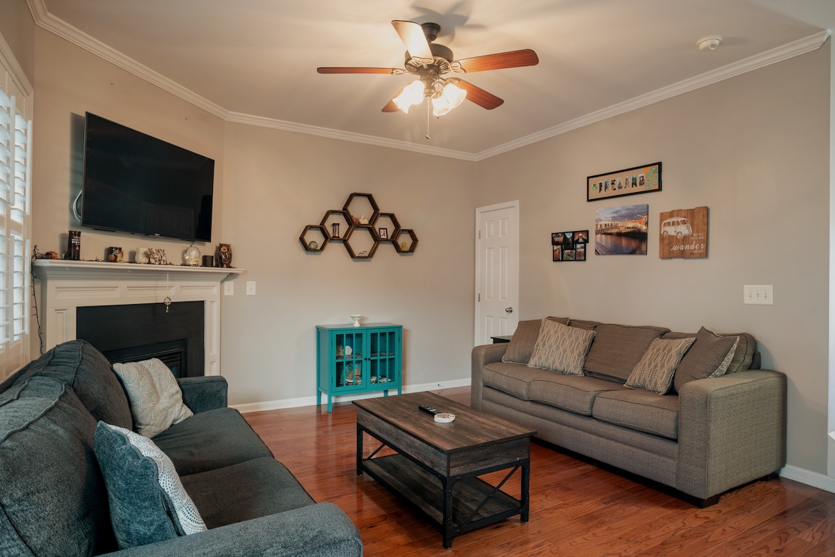 The living room features two gray sofas facing a wooden coffee table. A wall-mounted flat-screen TV is above an elegant fireplace. Light hardwood flooring complements the neutral wall colors. Decorative hexagonal frames are displayed on the wall, adding character to the space.