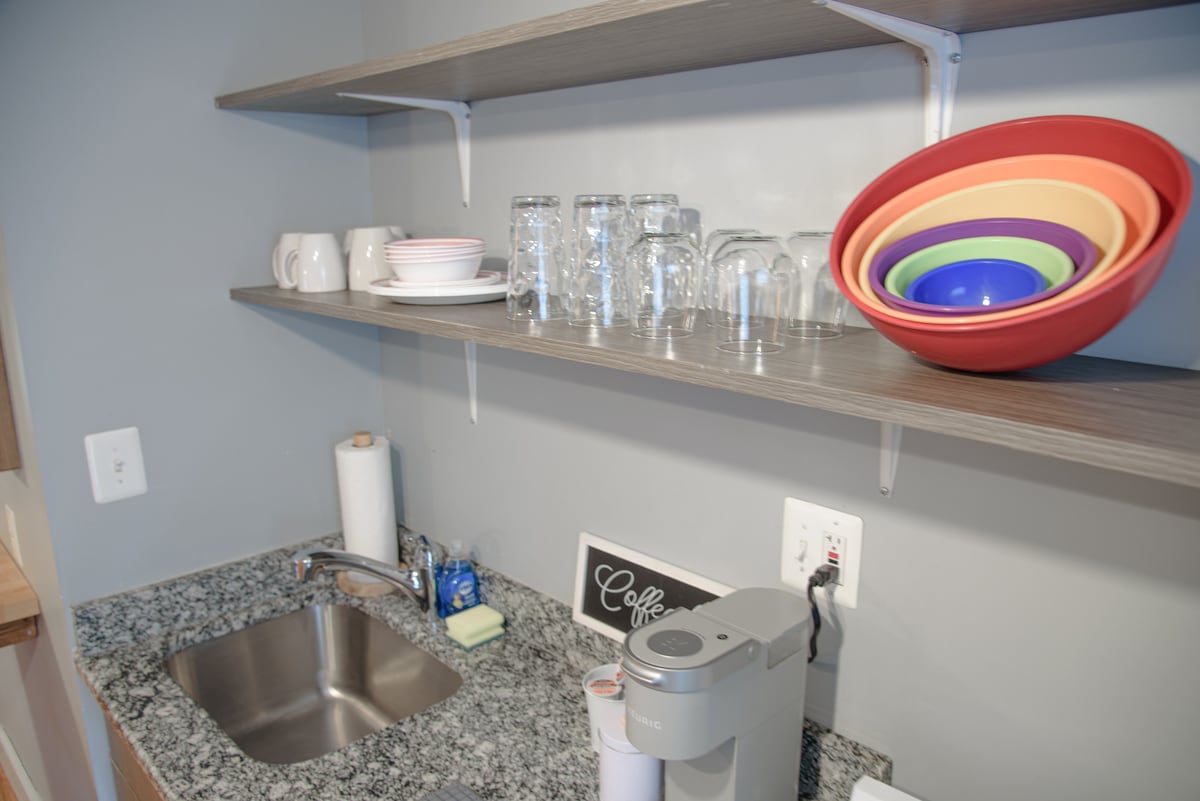 A kitchen space features open shelving displaying a variety of glassware, colorful mixing bowls, and dinnerware. A granite countertop houses a sink and a coffee maker, while a roll of paper towels is positioned nearby.