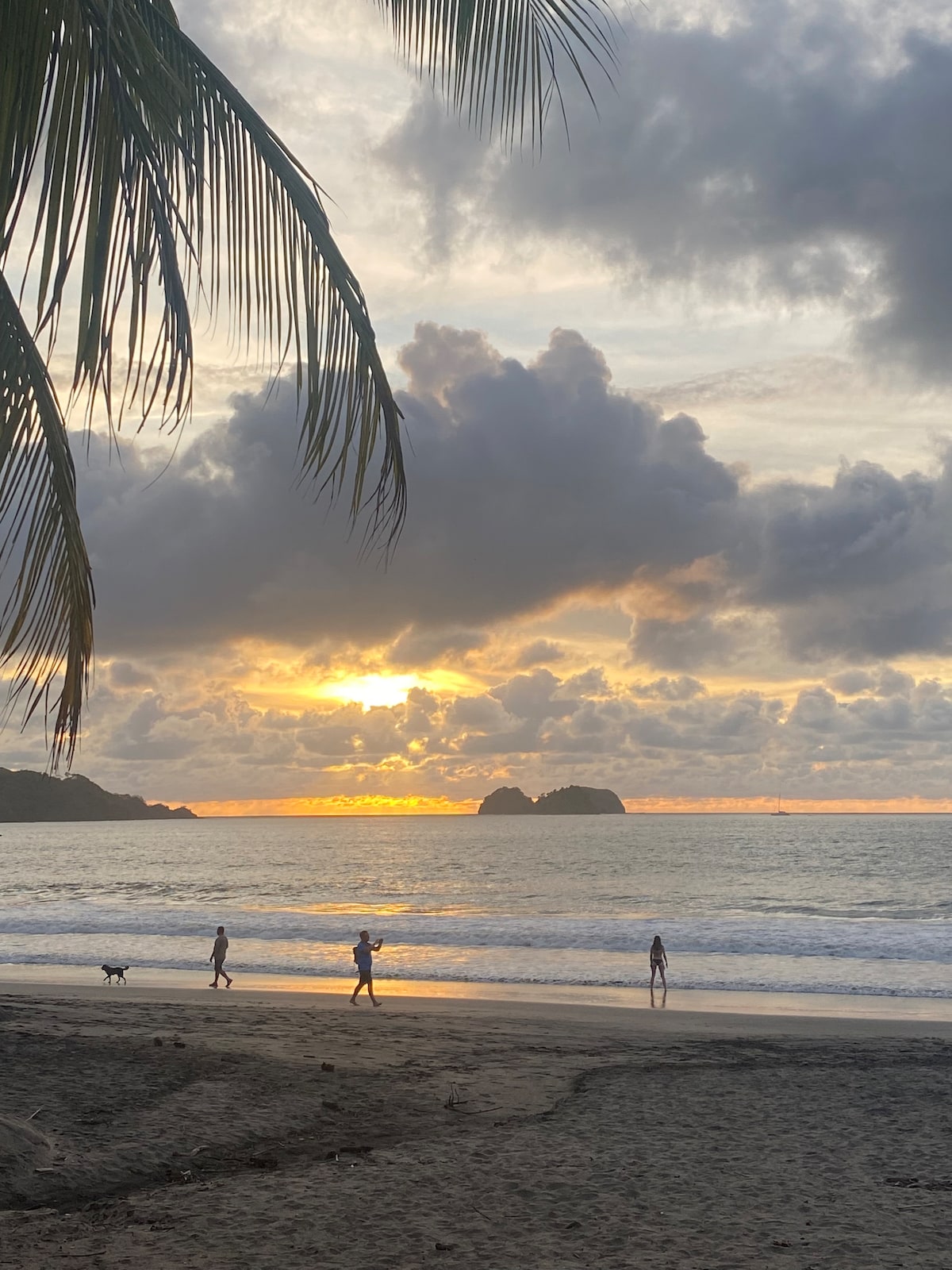 The image captures a serene beach scene at sunset, where silhouettes of people stroll along the shoreline. Gentle waves lap against the sand, and a distant rocky formation is visible near the horizon. Soft clouds scatter across the sky, reflecting warm hues of orange and yellow.