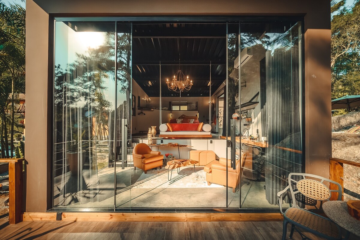 A modern living space is visible through large glass doors, showcasing a well-furnished area with an elegant chandelier and a cozy arrangement of orange chairs. A stylish sofa and wooden accents contribute to the overall ambiance, creating a bright and inviting indoor environment.