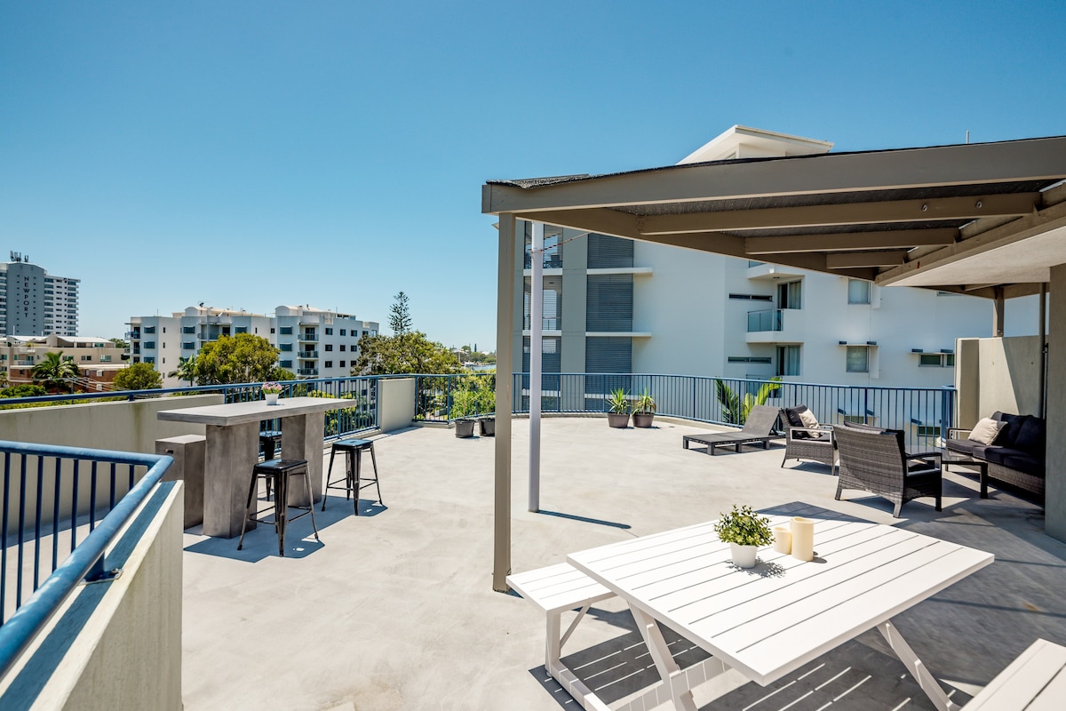 The private rooftop terrace offers a spacious lounge area with comfortable seating, a dining table, and a concrete bar, surrounded by views of Mooloolaba. The terrace features a clean, modern design with ample space for relaxing and enjoying outdoor gatherings.