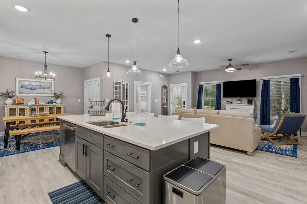 A modern kitchen is designed with an island featuring seating, accompanied by pendant lights. Adjacent, a cozy living area contains a comfortable sofa, a coffee table, and a mounted television, with large windows allowing natural light to fill the space. Decorative touches include a dining table and wall art.