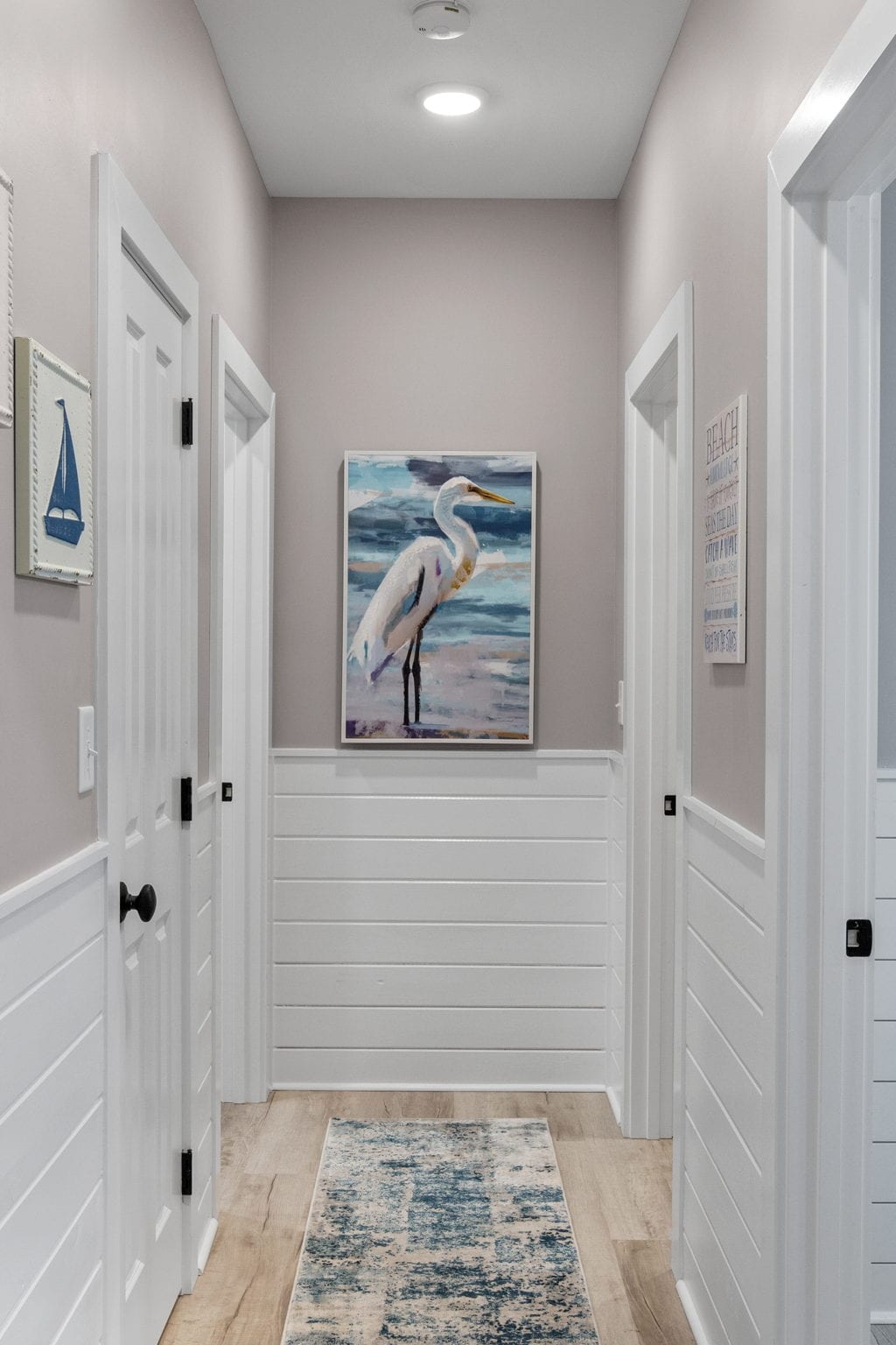 A well-lit hallway features light gray walls with white wainscoting. Two white doors are visible on either side, leading to private spaces. On the wall, a framed artwork of a heron adds a touch of color. A soft blue and white area rug runs along the wooden floor.