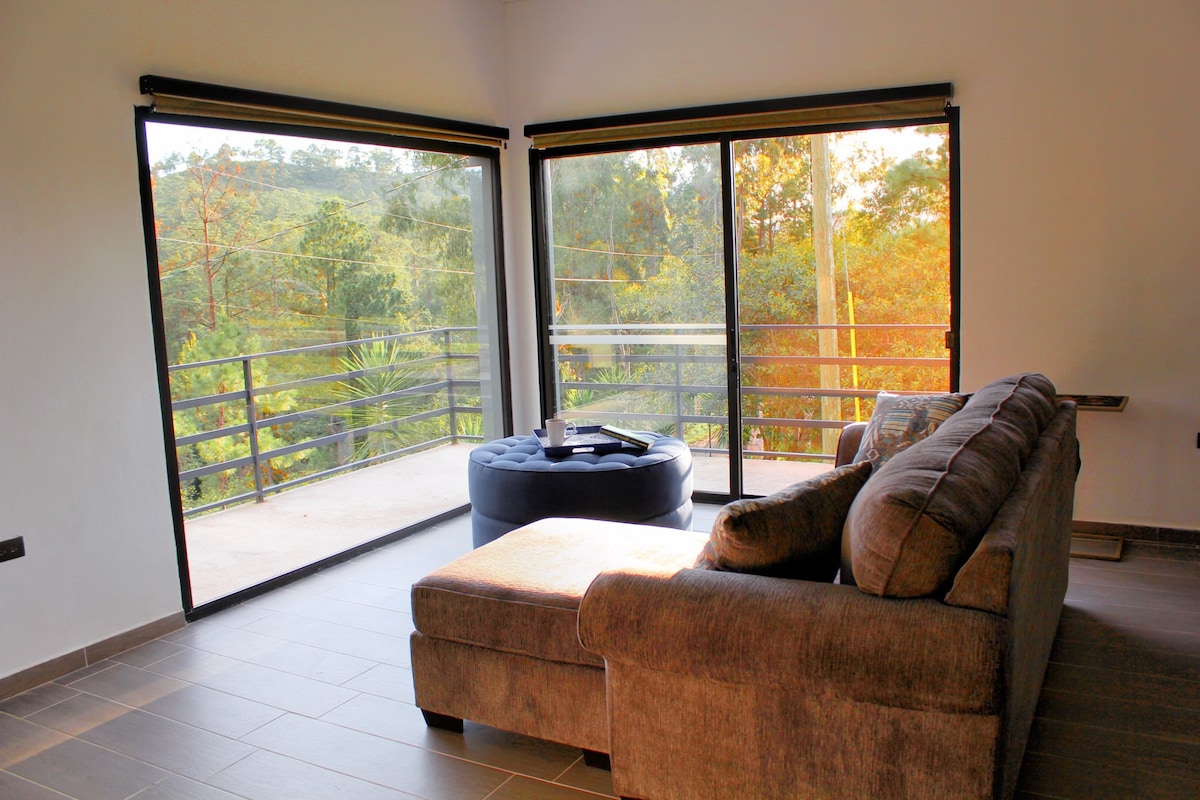 A spacious living area features large glass doors that open to a private balcony, showcasing views of the surrounding greenery. A comfortable sectional sofa and a round ottoman are visibly arranged for relaxation, with soft natural light filtering through the windows.
