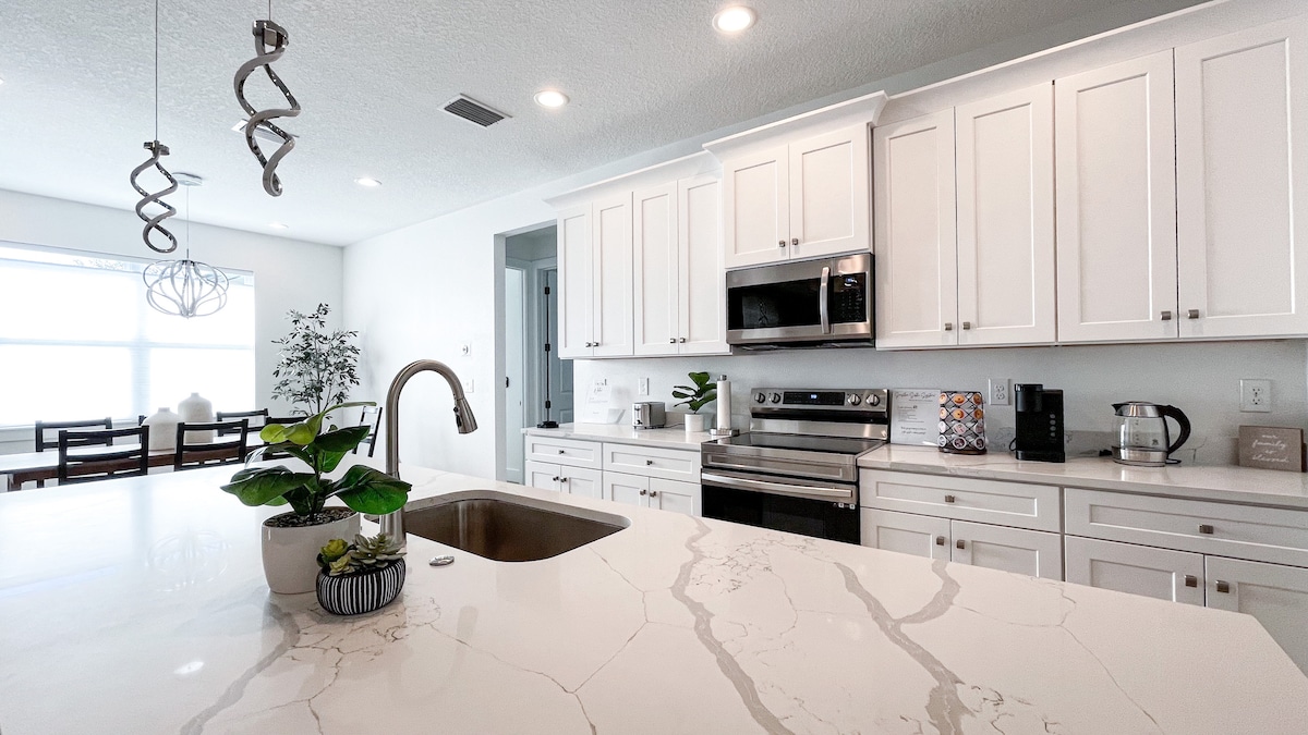 A modern kitchen features sleek white cabinetry and stainless steel appliances. A large marble countertop is accentuated by a central sink and a small potted plant. Natural light floods the space through a large window, enhancing the overall bright and inviting ambiance.