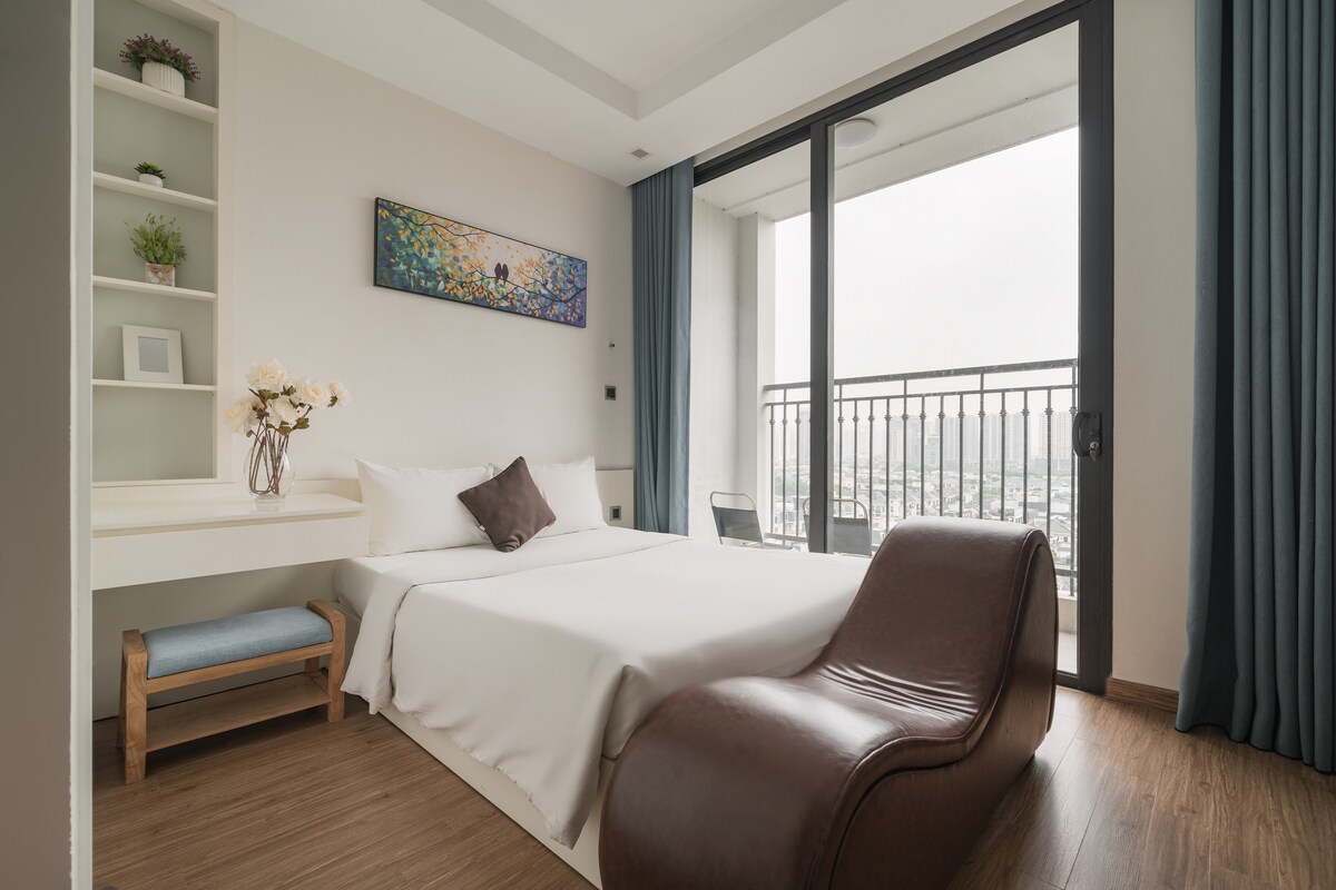 A neatly arranged bedroom features a spacious bed with crisp white linens. A comfortable chair sits beside a small writing desk, and large glass doors open to a balcony, allowing natural light to fill the room. City views are visible beyond the railing.