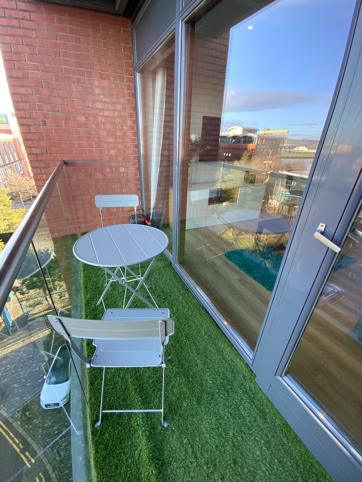 A small private balcony features a circular table and two folding chairs, set against a backdrop of glass panels. A green artificial lawn covers the floor, providing a cozy touch. Natural light brightens the space, enhancing views of the surrounding area.