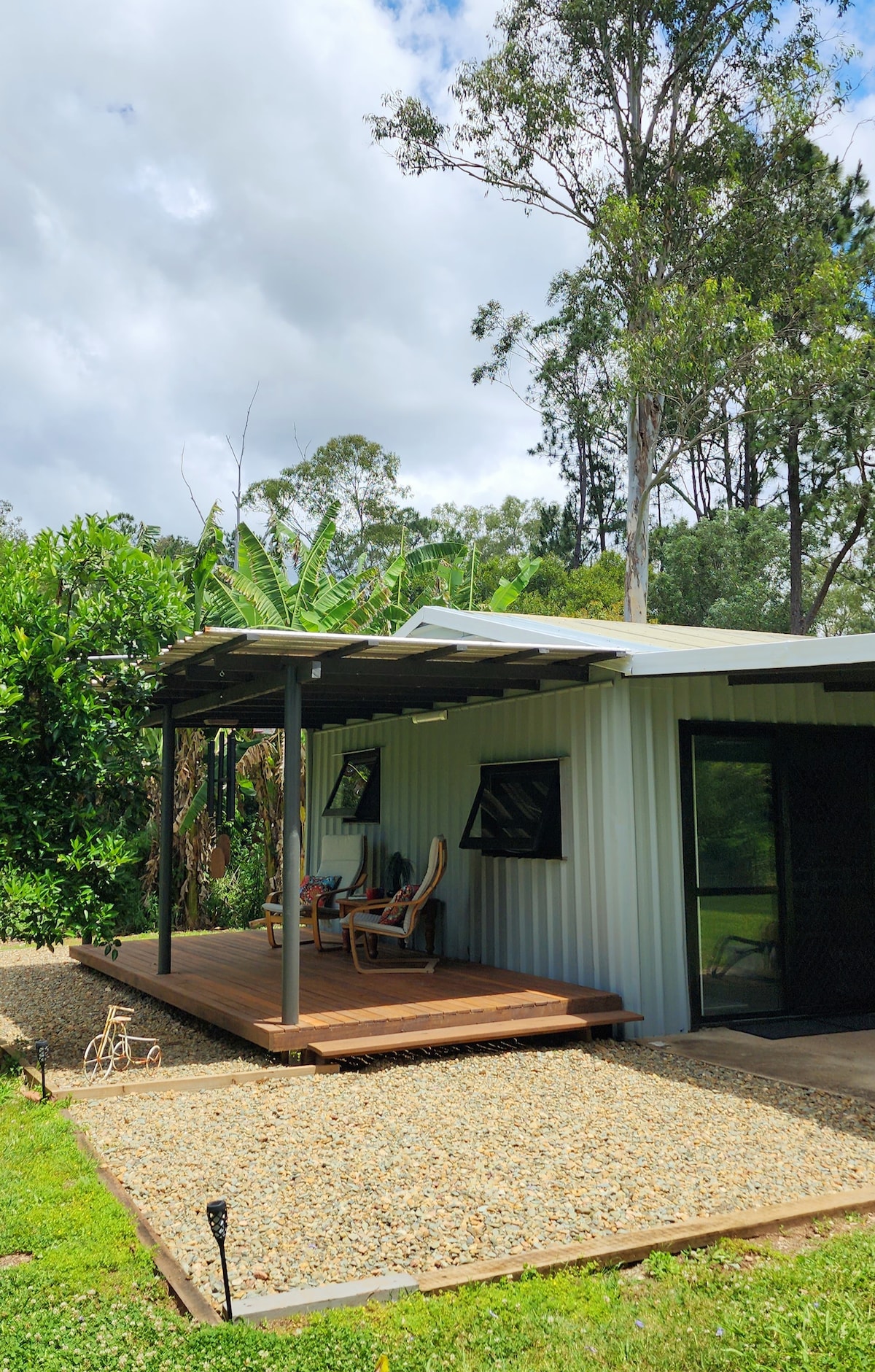 A cozy outdoor area is featured, with a wooden deck embraced by lush greenery. Two comfortable chairs are positioned under the covered space, perfect for relaxation. The exterior showcases a modern design with metallic siding and large windows, inviting natural light into the cabin.