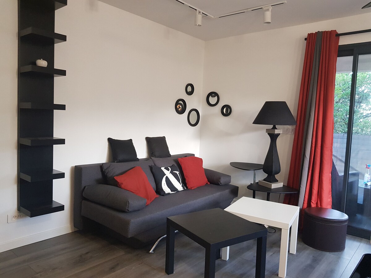 A modern living area is presented with a dark sectional sofa adorned with black and red cushions. A round side table and a small ottoman provide additional seating. A black lamp stands nearby, while wall-mounted shelves display decorative items. Large windows allow natural light to fill the space.